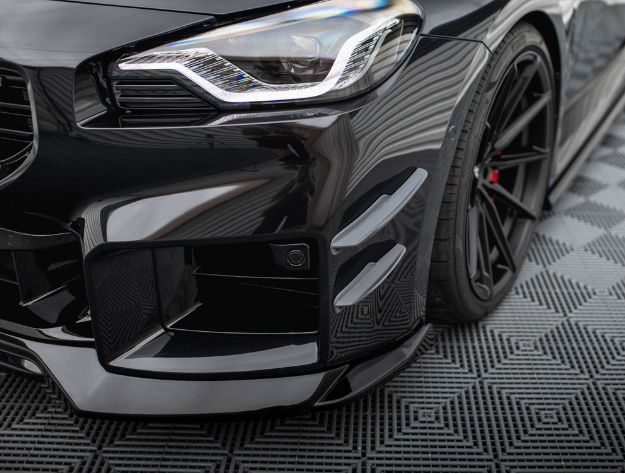 A sleek, black sports car shows its front headlight and grille, with a sporty bumper and aerodynamic features. It's parked on a textured, patterned surface, enhancing its modern appearance.