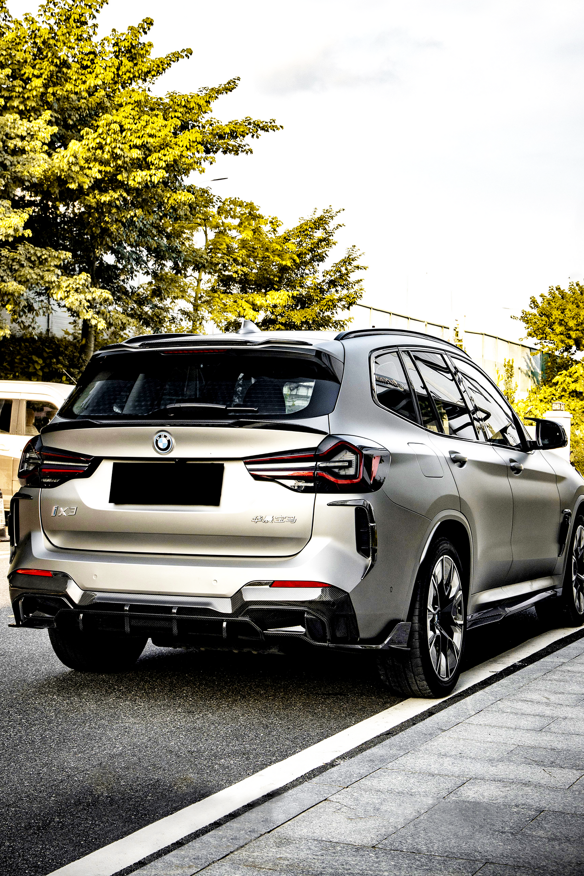 A silver SUV is parked on a road beside a sidewalk, surrounded by green trees. The vehicle has a BMW logo and "iX3" and "Electric" badges visible.