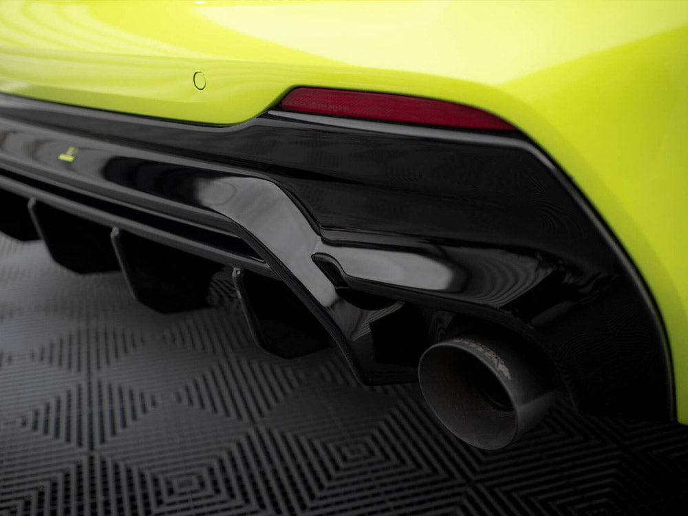A bright yellow sports car's rear bumper features a glossy black trim and a visible exhaust pipe, set on a textured, geometric-patterned floor.