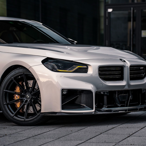 A silver car with black rims is parked on a paved surface. Its sleek design features modern headlights and a prominent grille, set against a dark urban backdrop.