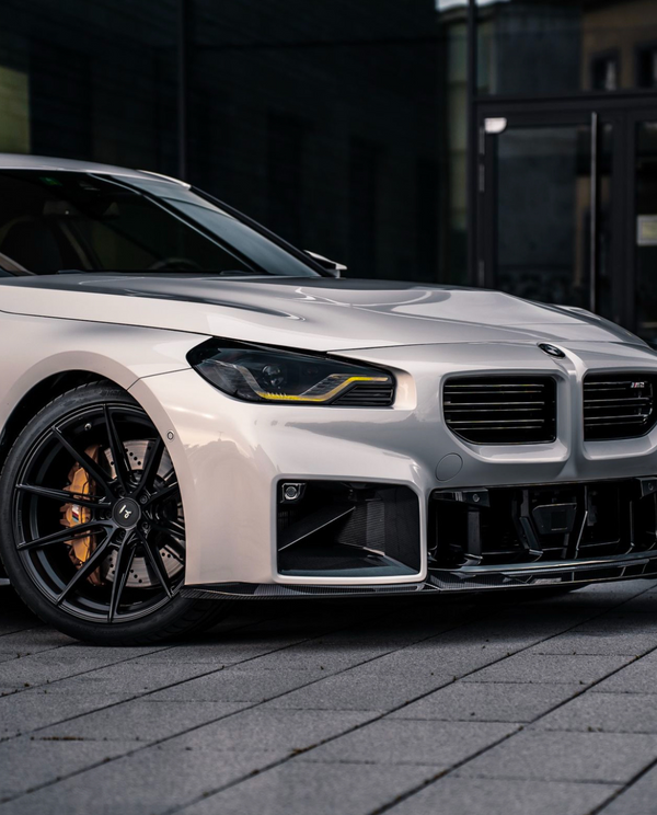 A silver car with black rims is parked on a paved surface. Its sleek design features modern headlights and a prominent grille, set against a dark urban backdrop.