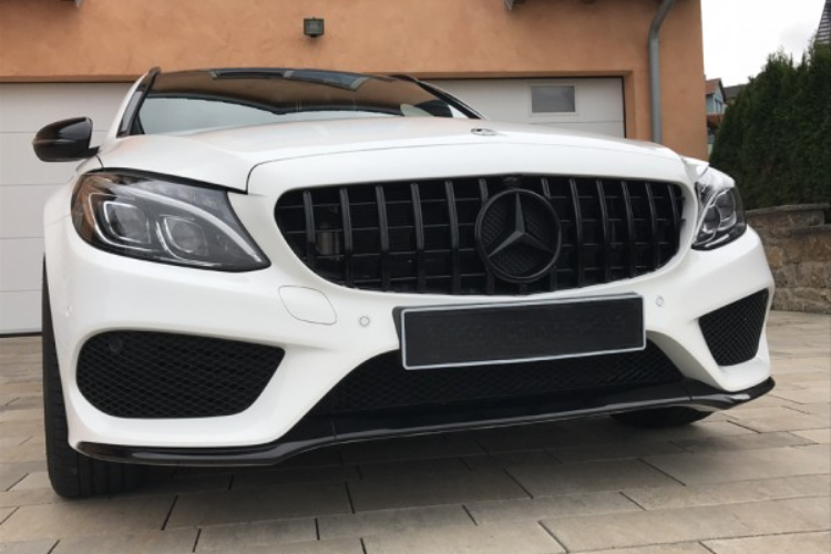 A white car with a black grille and Mercedes emblem is parked on a stone driveway in front of a garage. The house walls are a warm orange shade.