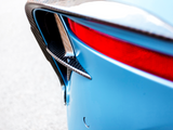 A car's rear end in close-up shows a carbon fiber vent, blue body, and red taillight. The scene is set on a light, neutral background.