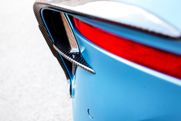 A car's rear end in close-up shows a carbon fiber vent, blue body, and red taillight. The scene is set on a light, neutral background.