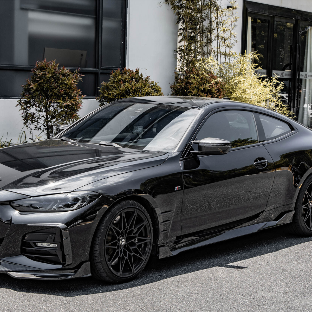 A sleek black sports car is parked beside modern office buildings, surrounded by neatly trimmed shrubs and plants, reflecting sunlight on its glossy surface.