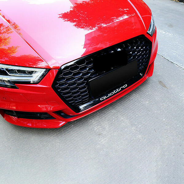 A red car displays a black honeycomb grille with "quattro" text below, parked on a concrete surface reflecting nearby trees.