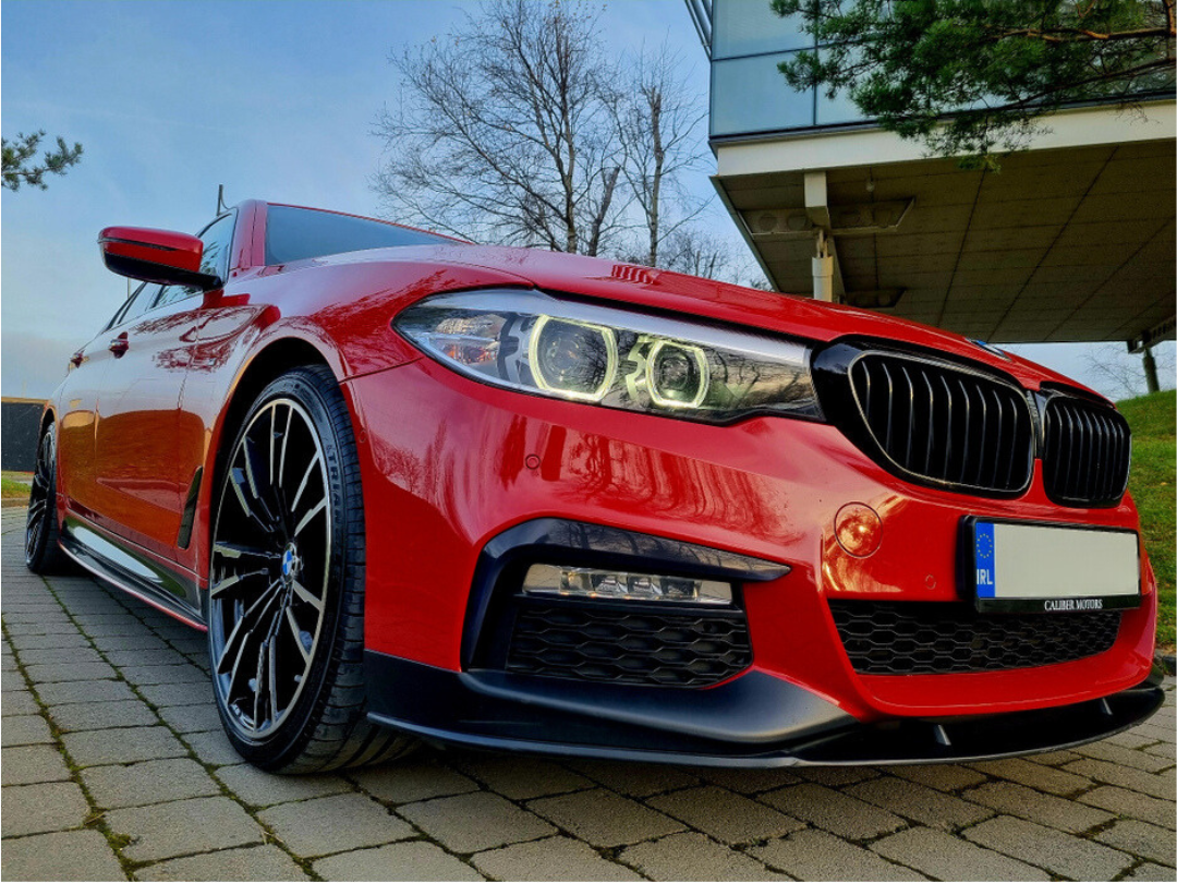 Red car parked on brick pavement, featuring sleek black rims and distinctive front grille; modern building and bare trees in background. License plate reads "IRL."