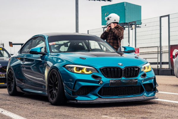A blue sports car with aggressive styling is parked on a racetrack. Its headlights are illuminated. Nearby, a person wearing a helmet stands, adjusting the strap in a fenced area.