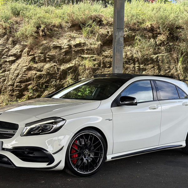 A white Mercedes-Benz car with AMG styling is parked under a structure. It is positioned on a dark paved surface, with a rocky, grassy hill in the background.