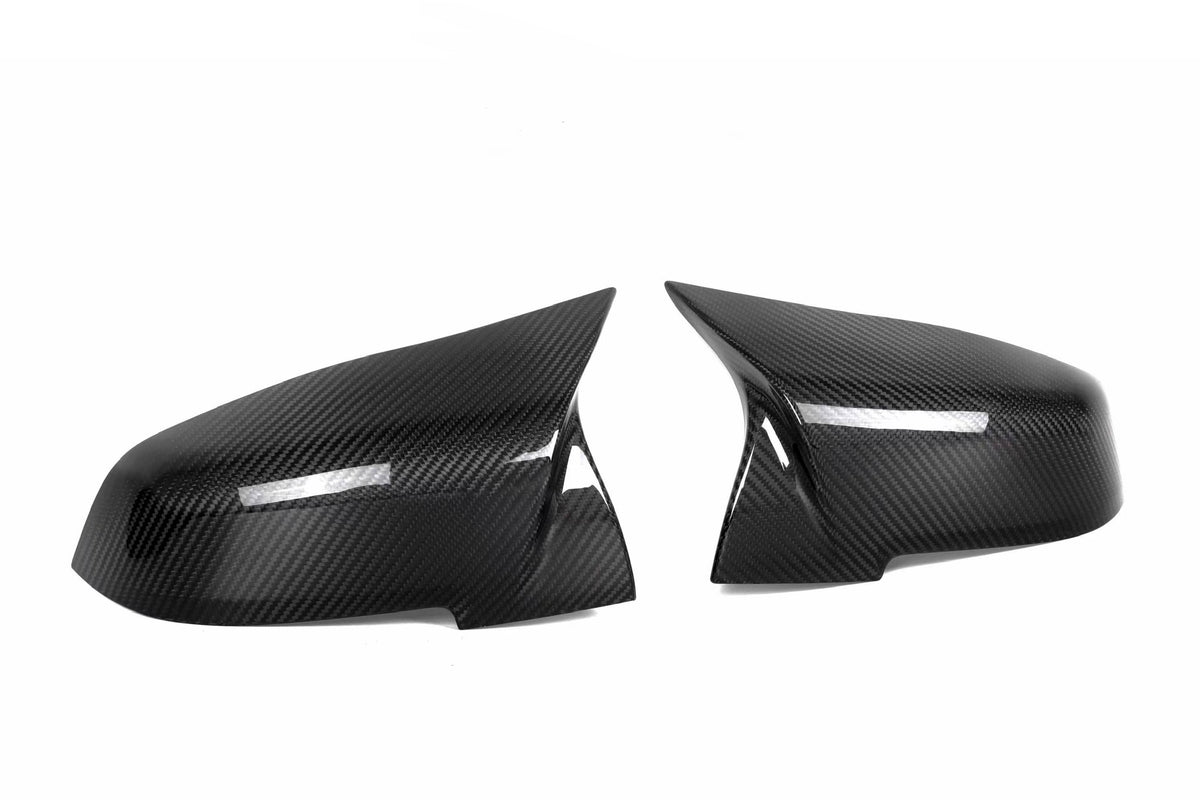 Two carbon fiber mirror covers are positioned side by side, facing each other against a plain white background, reflecting light subtly across their glossy, textured surfaces.