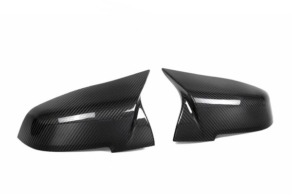 Two carbon fiber mirror covers are positioned side by side, facing each other against a plain white background, reflecting light subtly across their glossy, textured surfaces.
