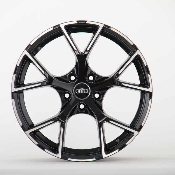 A glossy black car rim features five angular spokes and the Audi logo in the center, resting against a plain white background.