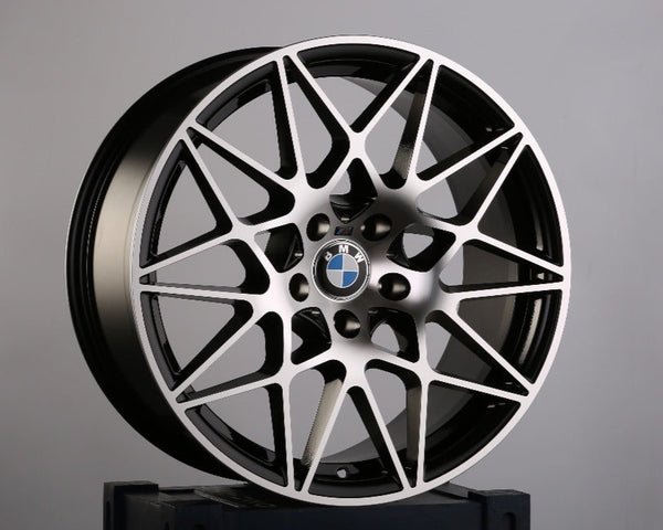 A black and silver BMW alloy wheel with intricate spoke design rests on a dark box. The BMW logo is visible at the center. The background is plain gray. Text on the box has "GA."
