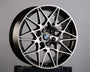 A black and silver BMW alloy wheel with intricate spoke design rests on a dark box. The BMW logo is visible at the center. The background is plain gray. Text on the box has "GA."