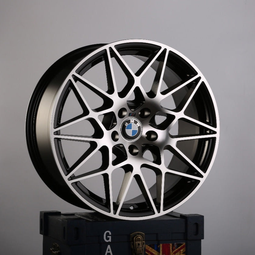 A black and silver BMW alloy wheel with intricate spoke design rests on a dark box. The BMW logo is visible at the center. The background is plain gray. Text on the box has "GA."