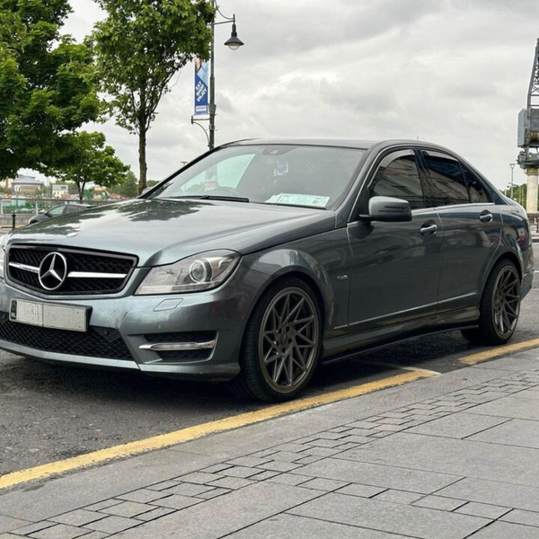A gray Mercedes sedan is parked on a city street near trees and a crane, under a cloudy sky. There is a license plate visible on the front of the car.