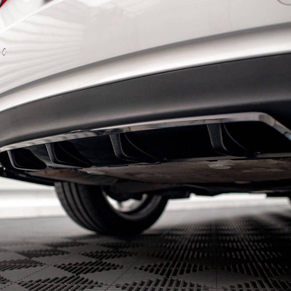 The rear of a white car with a black diffuser and dual exhaust pipes is parked on a checkered floor, against a backdrop of horizontal white panels.