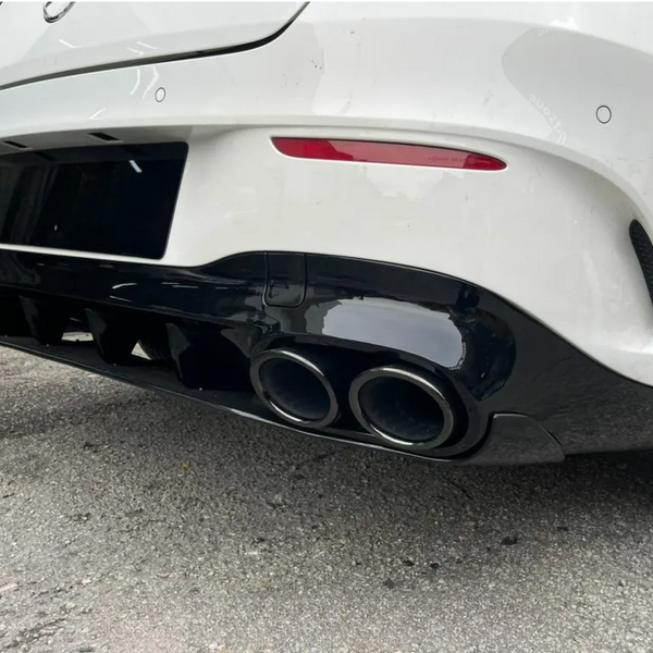 A car's rear bumper displays dual exhaust pipes, parked on a rough asphalt surface. The white vehicle features a black diffuser and a red reflector amid an urban environment.
