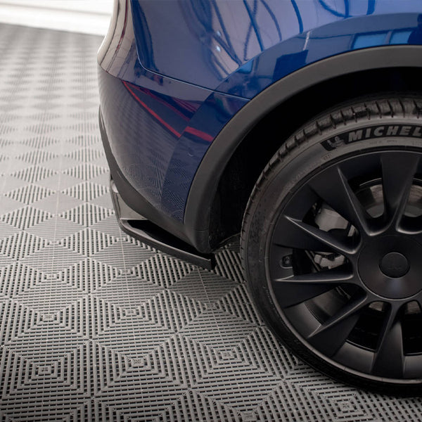 A car's rear bumper and black tire with "MICHELIN 255/40R20" text are parked on a patterned gray surface, reflecting a building's geometric outline.