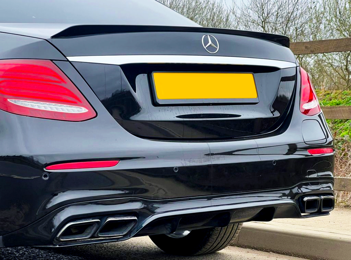 A black luxury car is parked by a wooden fence, showcasing its rear with red taillights and dual exhausts. The car has a yellow license plate and a silver emblem.