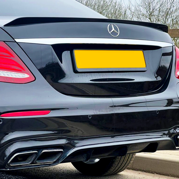 A black luxury car is parked by a wooden fence, showcasing its rear with red taillights and dual exhausts. The car has a yellow license plate and a silver emblem.