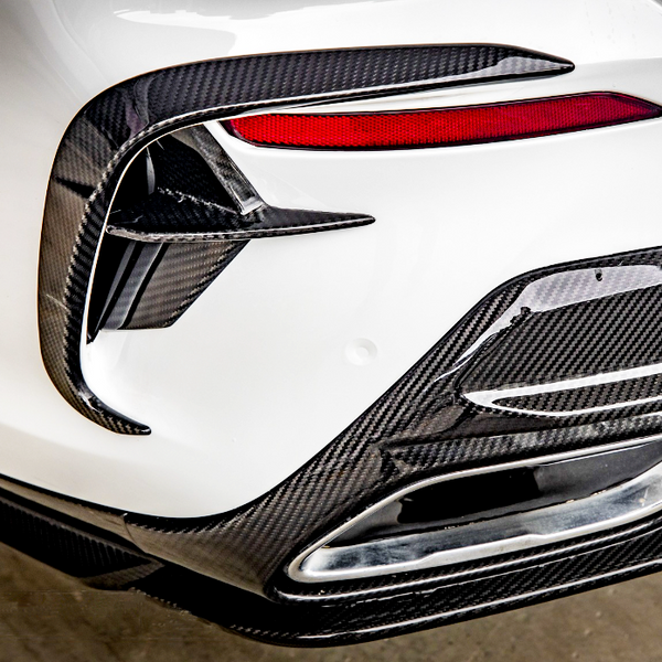 A car's rear bumper features carbon fiber detailing, red tail light accents, and an exhaust outlet, set against a clean, white body in a garage environment.