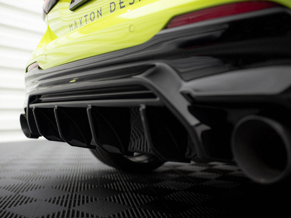 Car rear bumper with a black aerodynamic diffuser, showing multiple fins, in a garage setting with a textured floor. Text reads "MAXTON DESIGN" on the yellow car body.