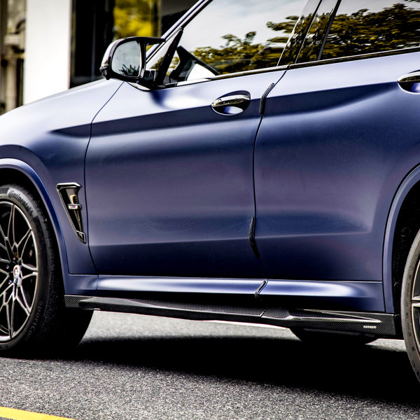 A sleek, dark blue car is parked on a street, showcasing its sporty design and black alloy wheels. Greenery and a white building wall form the background.