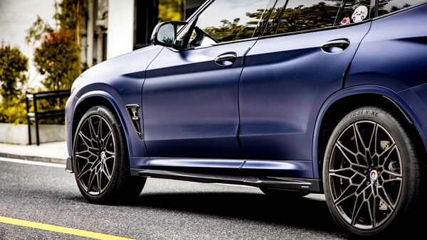A sleek, dark blue car is parked on a street, showcasing its sporty design and black alloy wheels. Greenery and a white building wall form the background.