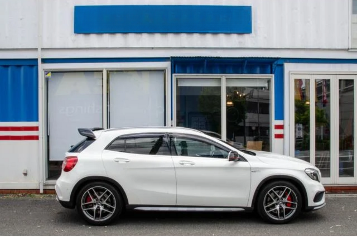 White car parked side view, in front of a building with glass doors and a blue and red striped facade. The car is stationary, and the setting is urban. No visible text.