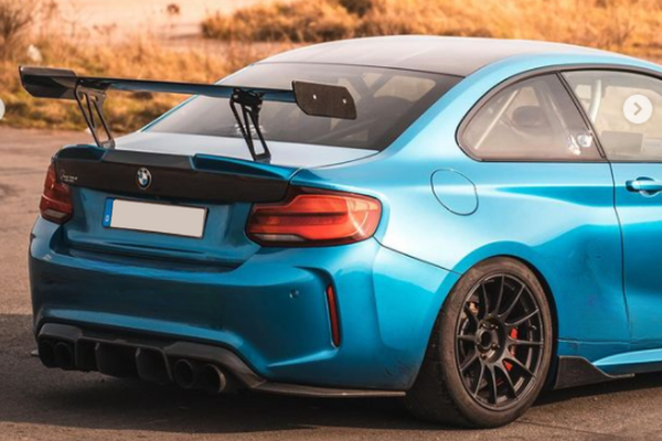 A blue sports car with a large rear wing is parked on a sunlit asphalt surface, surrounded by dry brown grass. The license plate area is blurred.