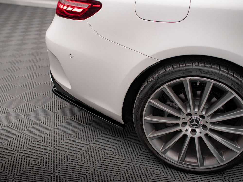 A car's rear end with a lit taillight, parked, showcases a silver alloy wheel and tire on a textured gray surface. The Mercedes-Benz logo is visible at the wheel's center.