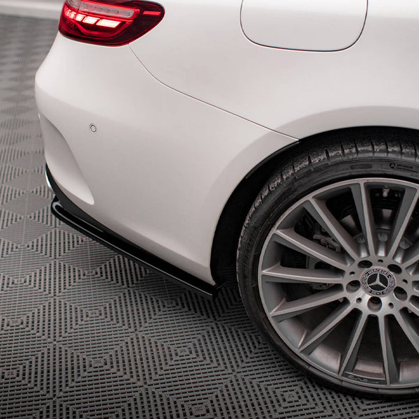 A car's rear end with a lit taillight, parked, showcases a silver alloy wheel and tire on a textured gray surface. The Mercedes-Benz logo is visible at the wheel's center.