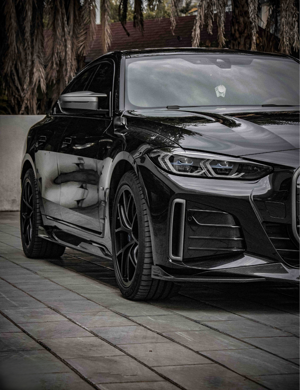 A sleek black car, parked on a tiled surface, reflecting surrounding trees and buildings under a cloudy sky, with palm leaves visible in the background.