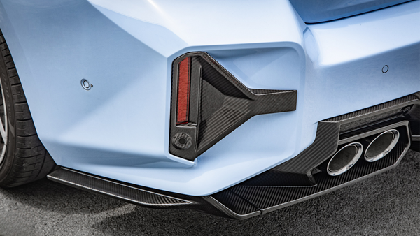 A light blue car's rear, featuring a carbon fiber diffuser and red taillight, with dual exhaust pipes and visible tire tread, on a textured road surface.