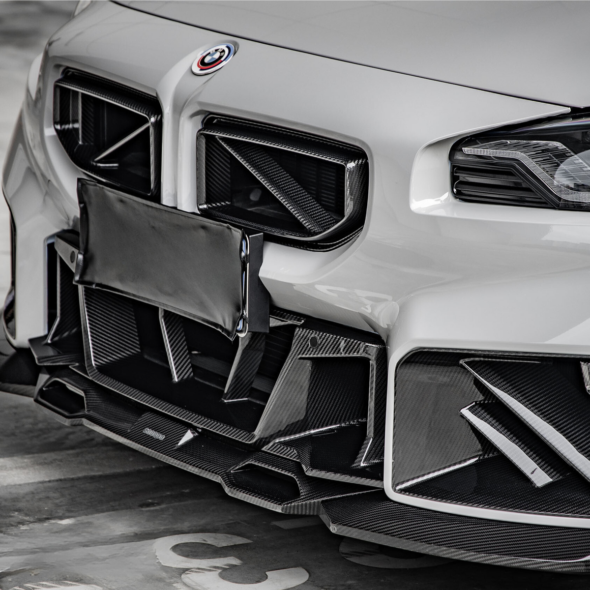 A white car with a sporty front bumper featuring intricate carbon fiber accents, parked in a concrete garage. A covered license plate is visible in the center.