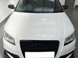 A white Audi car is parked in a garage, viewed from the front, displaying its black grille with the Audi logo prominently in the center. The environment is industrial.