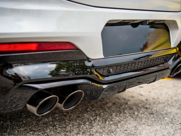The car's rear features a sleek black bumper with dual exhaust pipes, framed by a white body and red tail light, parked on a gray asphalt surface beside another vehicle.