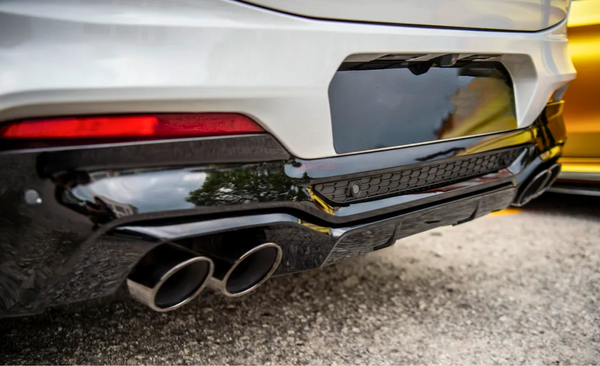 The car's rear features a sleek black bumper with dual exhaust pipes, framed by a white body and red tail light, parked on a gray asphalt surface beside another vehicle.