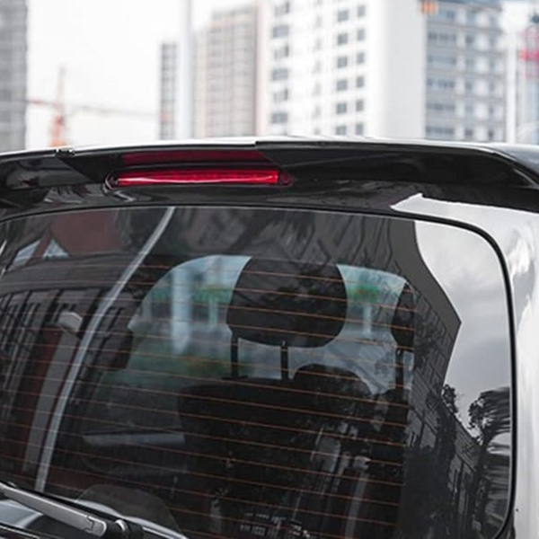 A black vehicle's rear window reflects surrounding trees and buildings, with a red brake light on top. In the background, urban buildings and construction cranes are visible.