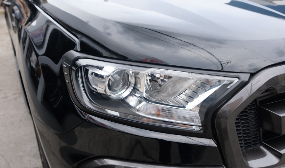 A sleek black vehicle's headlight assembly, reflecting light, is prominently displayed. It is parked on a concrete surface, with a cloudy sky reflecting on the car's surface.