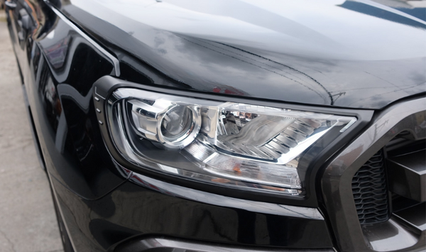 A sleek black vehicle's headlight assembly, reflecting light, is prominently displayed. It is parked on a concrete surface, with a cloudy sky reflecting on the car's surface.