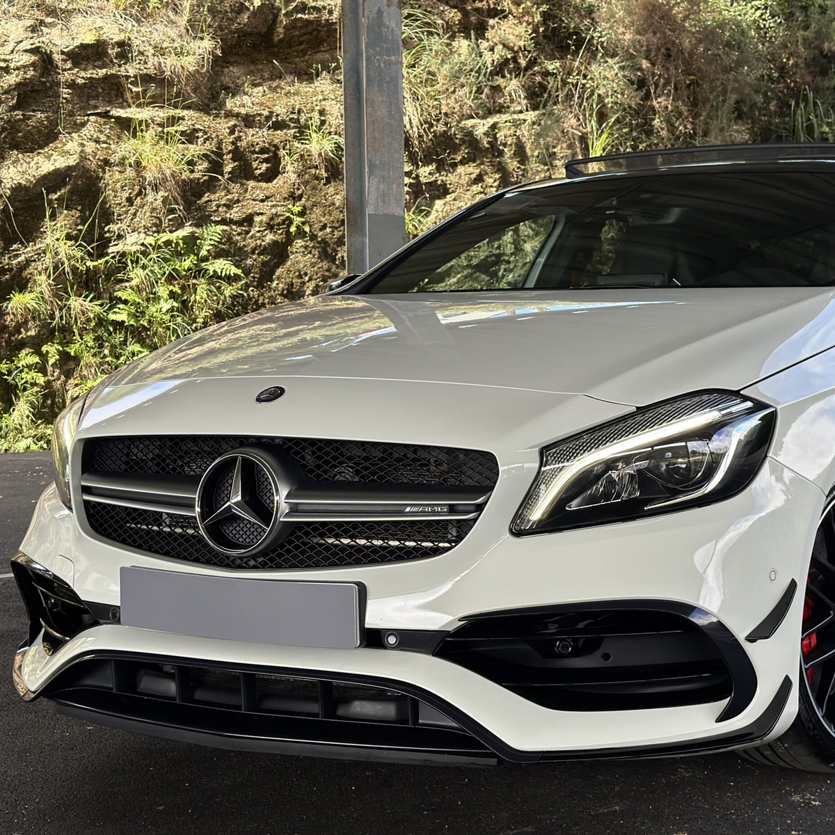 White Mercedes-Benz car sits parked under a metal structure; its front grille features the Mercedes logo and “AMG” badge; lush greenery and rocky terrain are visible in the background.