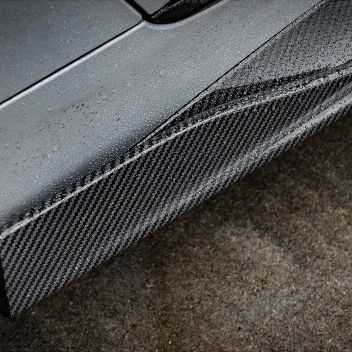 A sleek car's carbon fiber side skirt, covered in raindrops, sits adjacent to a wet asphalt surface, highlighting the vehicle's modern design and performance-oriented components.