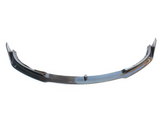 A black carbon fiber car front lip spoiler rests on a plain white background, showcasing its sleek, aerodynamic design with raised ends and slight curvature.