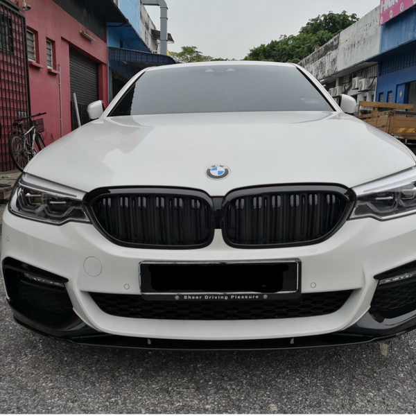 A white BMW car is parked on a gravel surface between industrial buildings. Text on the license plate area reads "Sheer Driving Pleasure." A bicycle and wooden pallets are nearby.