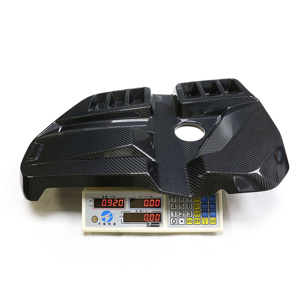 A carbon fiber engine cover rests on a digital scale, displaying measurements: "0.920" and "1.00". The background is a plain white surface.