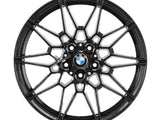 A black, intricately designed alloy wheel rim with the BMW logo at its center, viewed directly from the front, set against a plain white background.