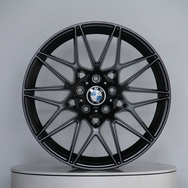 A black BMW alloy wheel with intricate spoke design is displayed vertically on a circular platform. The center features the BMW logo. The background is plain and light gray.