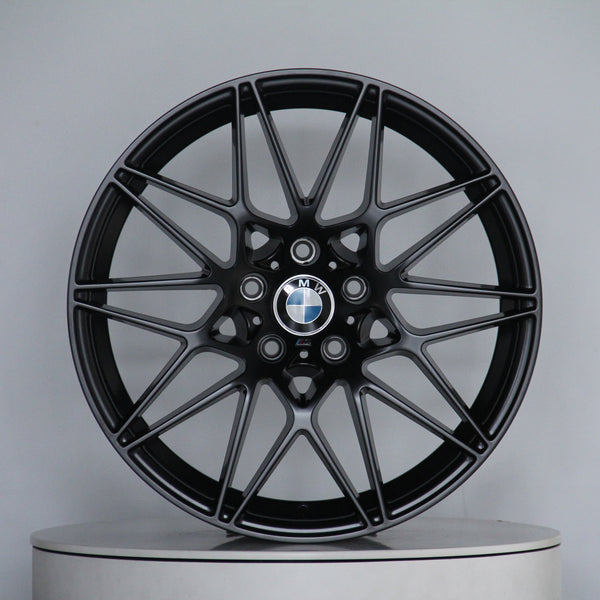 A sleek black alloy wheel with a multi-spoke design, featuring the BMW logo at its center, stands on a plain circular platform in a light gray setting.